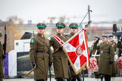 Poczet sztandarowy Kompanii Reprezentacyjnej Straży Granicznej idzie po placu Marszałka Józefa Piłsudskiego w Warszawie. W tle funkcjonariusz oddający honor sztandarowi i orkiestra wojskowa.