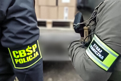 Rękawy stroju dwóch funkcjonariuszy z opaską z napisem CBŚP Policja oraz Straż Graniczna. W tle rozmazane palety z kartonami.