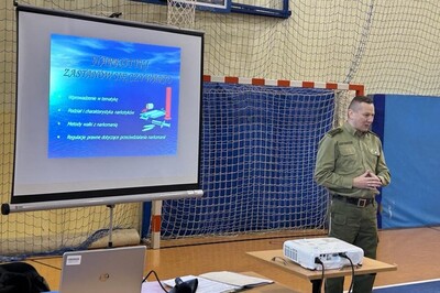 Prelekcje funkcjonariusza z PSG w Zakopanem Funkcjonariusz Straży Granicznej przemawiający do uczniów podczas prelekcji. obok niego telebim z informacjami dotyczącymi tematu prelekcji.