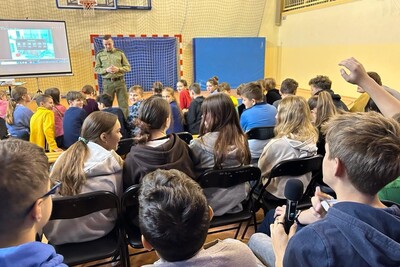 Prelekcje funkcjonariusza z PSG w Zakopanem Funkcjonariusz Straży Granicznej przemawiający do uczniów podczas prelekcji. za nim telebim z informacjami dotyczącymi tematu prelekcji. uczniowie siedzą na ławkach. wydarzenie odbywa się na sali gimnastycznej. uczniowie zdają pytania funkcjonariuszowi.