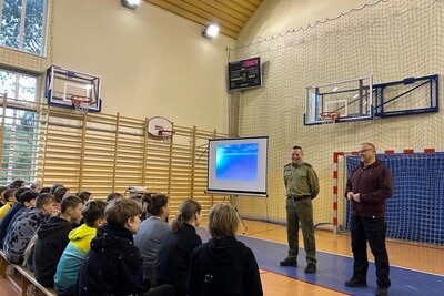 Prelekcje funkcjonariusza z PSG w Zakopanem Funkcjonariusz Straży Granicznej przemawiający do uczniów podczas prelekcji. obok niego z lewej strony telebim z informacjami dotyczącymi tematu prelekcji natomiast z prawej strony stoi prawdopodobnie nauczyciel lub dyrektor szkoły. uczniowie siedzą na ławkach. wydarzenie odbywa się na sali gimnastycznej.
