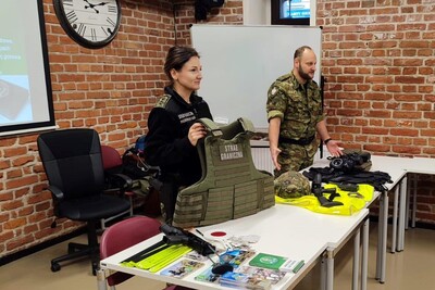 Sala wykładowa.  Funkcjonariuszka i funkcjonariusz SG stoją za stołem, na którym leżą ulotki informacyjne, kamizelki SG, hełm, lornetka i kajdanki. Funkcjonariuszka trzyma w rękach kamizelkę taktyczną z napisem Straż Graniczna.