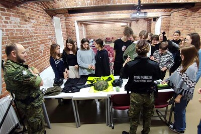 Sala wykładowa. Funkcjonariuszka i funkcjonariusz SG stoją tyłem. Przed nimi stół, na którym leżą ulotki informacyjne, kamizelki SG, hełm, lornetka i kajdanki. W tle stoją uczniowie uczestniczący w spotkaniu.