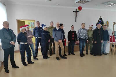 Aula. Grupa odwiedzających placówkę z okazji mikołajek. W grupie są niepełnosprawne osoby dorosłe oraz ich opiekunowie. W półkolu stoją: ksiądz biskup, funkcjonariusze SG, policjanci, mężczyzna przebrany za św. Mikołaja - biskupa.