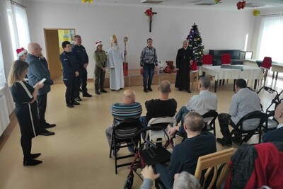 Aula. Grupa około 30  uczestników spotkania z biskupem, funkcjonariuszami SG i policjantami z okazji mikołajek. W grupie są niepełnosprawne osoby dorosłe oraz ich opiekunowie. Oglądają występ. Przemawia ksiądz biskup. Za nim stoją funkcjonariusze SG, policjanci, mężczyzna przebrany za św. Mikołaja - biskupa.