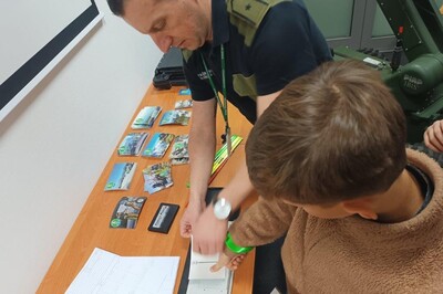 Pomieszczenia Placówki SG w Krakowie – Balicach. Chłopiec stoi przy biurku. Funkcjonariusz SG odbija w tuszu jego kciuk – pobiera odcisk palca. Na biurku leżą widokówki i odblaski, promujące KaOSG.
