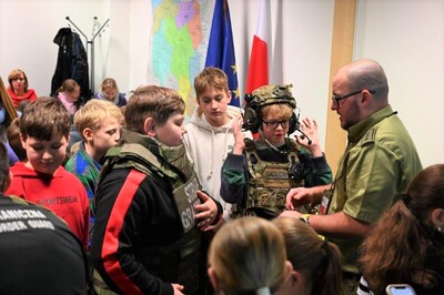 Pomieszczenia Placówki SG w Krakowie – Balicach. Grupa młodzieży około 15 osobowa stoi przy funkcjonariuszu SG, który prezentuje sprzęt używany na co dzień przez SG . Jeden chłopiec ma założoną kamizelkę kuloodporną, a drugi kamizelkę taktyczną oraz hełm ze słuchawkami i mikrofonem.