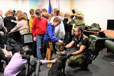 Pomieszczenia Placówki SG w Krakowie – Balicach. Grupa młodzieży około 20 osobowa stoi przy funkcjonariuszach SG. Jeden z nich prezentuje sprzęt używany na co dzień przez SG i pomaga ubrać chłopcu kamizelkę taktyczną. Drugi funkcjonariusz kusa, trzymając psa służbowego. Chłopiec i dziewczynka głaskają psa.