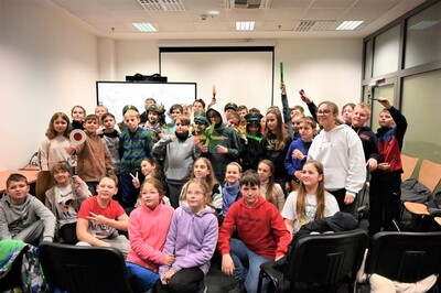 Pomieszczenia Placówki SG w Krakowie – Balicach. Grupa ponad 40 dzieci stoi i uśmiecha się do zdjęcia. Dzieci trzymają w rękach odblaski, lizak do zatrzymywania pojazdów. Kilkoro dzieci ma na głowach czapki służbowe, hełm.