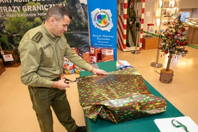 Sala Tradycji KaOSG funkcjonariusz SG stoi przy stole i pakuje w ozdobny kolorowy papier prezent. W tle kilka spakowanych już prezentów, choinka oraz baner z logotypem i napisem Związek Zawodowy pracowników ministerstwa spraw wewnętrznych oraz eksponaty muzealne, które na stałe znajdują się w Sali tradycji.