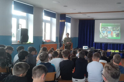 Aula szkoły. Funkcjonariuszka stoi przed kilkudziesięcioosobowa grupą młodzieży, prowadzi prelekcję na temat służby w Straży Granicznej. W tle ekran z prezentacja o Straży Granicznej.