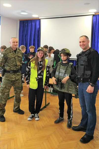 Aula szkoły. Umundurowany funkcjonariusz SG stoi z dziewczynką, chłopcem i nauczycielem. Dzieci są ubrane w sprzęt wykorzystywany na co dzień w służbie przez funkcjonariuszy kamizelkę taktyczną, kamizelkę odblaskową, czapkę, hełm. Chłopiec trzyma w dłoniach lornetkę. Nauczyciel ma ubraną czarną kamizelkę taktyczną.