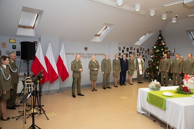 Spotkania wigilijne w Karpackim Oddziale Straży Granicznej komendant karpackiego oddziału przemawia do zebranych na spotkaniu  wigilijnym  funkcjonariuszy oraz pracowników karpackiego oddziału straży granicznej. w tle choinka. przed komendantem stoi stół na którym znajdują się stroik wigilijny oraz opłatki.