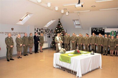 Spotkania wigilijne w Karpackim Oddziale Straży Granicznej kapelan karpackiego oddziału straży granicznej odmawia modlitwę do zebranych na spotkaniu  wigilijnym  funkcjonariuszy oraz pracowników karpackiego oddziału straży granicznej. w tle choinka. przed kaplanem stoi stół na którym znajdują się stroik wigilijny oraz opłatki.