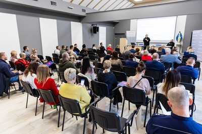 Sala wykładowe. Funkcjonariuszka SG stoi przed uczestnikami konferencji i przemawia do mikrofonu. Przy stole siedzi funkcjonariusz SG. Sala wypełniona jest siedzącymi słuchaczami prelekcji.