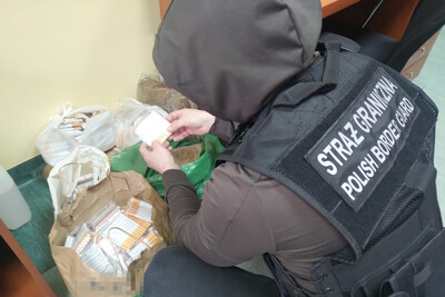 Funkcjonariusz w ubraniach cywilnych z założoną czarną kamizelką taktyczną z napisem Straż Graniczna Polish Border Guard ogląda zatrzymany towar. Trzyma w ręce papierosy. Na ziemi leży kilka toreb wypełnionych papierosami.