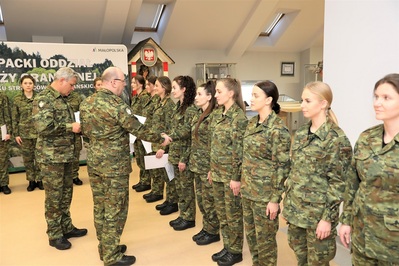 Sala Tradycji KaOSG. W szeregu stoi  9 funkcjonariuszek SG w umundurowaniu polowym. Pułkownik  Marcisz wręcza akt mianowania jednej z funkcjonariuszek SG. Towarzyszy mu naczelnik wydziału kadr i i szkolenia, który podaje dokumenty.