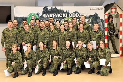 Sala Tradycji KaOSG. W dwóch szeregach stoi 19 uśmiechniętych funkcjonariuszy SG w umundurowaniu polowym, w kolejnym pierwszym rzędzie 8 funkcjonariuszek przykucnęło przed resztą, by ładnie zapozować do zdjęcia grupowego. Wszyscy w rekach trzymają akty mianowania na stopień kaprala SG.