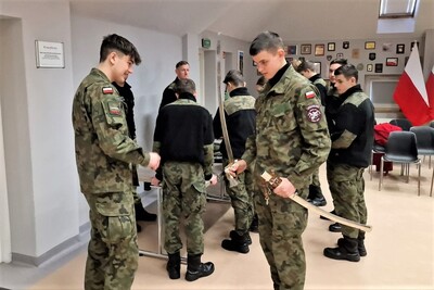Sala tradycji. Uczniowie z klasy mundurowej zapoznają się ze sprzętem służbowym Straży Granicznej. Jeden z młodych mężczyzn trzyma w dłoni szablę. W tle kilku innych uczniów ogląda sprzęt służbowy pod okiem funkcjonariusza KaOSG.