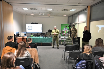 Sala szkoleniowa w siedzibie Placówki SG w Krakowie-Balicach. Funkcjonariusz SG stoi przed studentami. Opowiada o służbie w Straży Granicznej. Na sali wystawiony jest sprzęt służbowy używany na co dzień przez funkcjonariuszy SG, stoi manekin w umundurowaniu specjalnym SG oraz robot pirotechniczny.