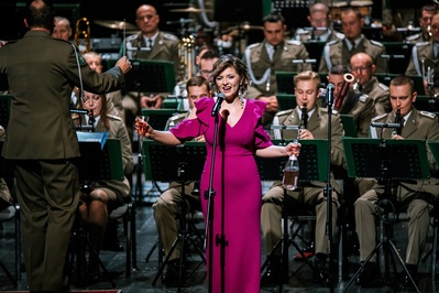 fot. Kinga Ptak, archiwum Małopolskiego Centrum Kultury SOKÓŁ na pierwszym planie solistka orkiestry reprezentacyjnej straży granicznej, występująca w długiej różowej sukni, wykonująca utwór podczas koncertu. na drugim planie muzycy orkiestry.