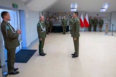 Sala Tradycji karpackiego Oddziału SG. Generał brygady Laciuga przyjmuje meldunek od prowadzącego uroczystość. W tle stoją zastępcy komendanta, kapelan oraz funkcjonariusze KaOSG. Obok nich funkcjonariusz orkiestry reprezentacyjnej  z trąbką.