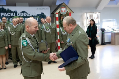Sala tradycji karpackiego oddziału sg naczelnik wydziału granicznego żegna swojego funkcjonariusza. Wręcza mu okolicznościowe prezenty. W tle funkcjonariusze uczestniczący w pożegnaniu i żona emeryta