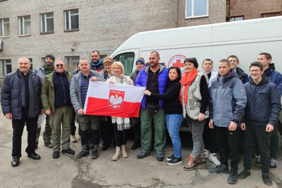 Wolontariusze i obdarowani stoją w grupie, kilkoro trzyma polską flagę, za nimi bus i budynek.