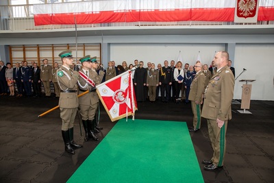 Komendant odchodzący na emeryturę i komendant obejmujący stanowisko Komendanta Karpackiego Oddziału Straży Granicznej stojący przed Sztandarem Karpackiego Oddziału Straży Granicznej. Na drugim planie stoją zaproszeni goście i przyglądają się przekazaniu sztandaru nowemu Komendantowi.