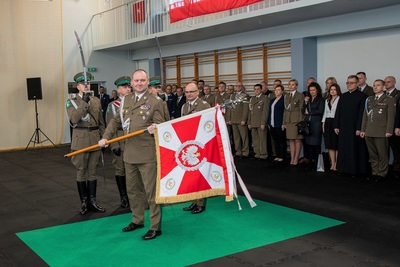 Komendant obejmujący stanowisko Komendanta Karpackiego Oddziału Straży Granicznej trzymający w dłoniach i prezentujący  Sztandar Karpackiego Oddziału Straży Granicznej. Na drugim planie stoją zaproszeni goście.