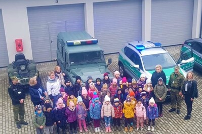 Na placu przed placówka sg w tarnowie dzieci stoja w zwartej grupie i pozują do zdjęcia na tle oznakowanych samochodów służbowych sg i garaży placówki sg w tarnowie.