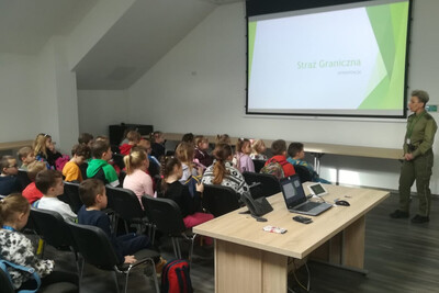 sala odpraw placówki sg w tarnowie. grupa dzieci siedzi przed ekranem na którym wyświetlana jest prezentacja. funkcjonariuszka sg opowiada im o historii formacji