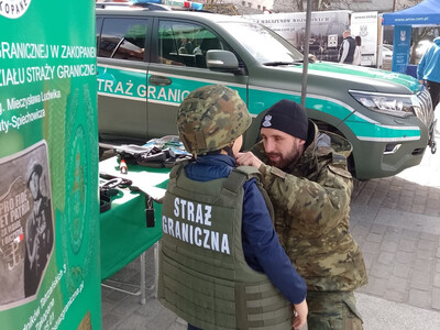 funkcjonariusz placówki sg ww zakopanem pomaga przymierzyć chłopcu kamizelkę taktyczną sg i hełm. w tle stanowisko promocyjne sg i samochód służbowy