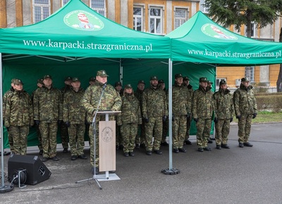 Plac apelowy w komendzie karpackiego oddziału straży granicznej. Komendant kaosg przemawia przy mównicy. Za nim pod namiotem  stoi kadra kierownicza oddziału