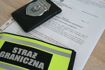 Na biurku leży decyzja o zobowiązaniu cudzoziemca do powrotu. Na niej odznaka funkcjonariusza sg i opaska zakładana na ramię z napisem straż graniczna