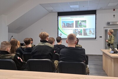 sala odpraw w tarnowskiej placówce sg grupa uczniów w mundurach siedzi tyłem na krzesłach, ogląda prezentację. Po prawej stronie stoi funkcjonariuszka będąca narratorem prezentacji. Na ekranie zdjęcia funkcjonariuszy sg podczas służby