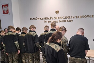 uczniowie stoją tyłem przy stole i przyglądają się rozłożonemu na nim sprzętowi służbowemu  w tle na ścianie napis placówka straży granicznej w Tarnowie im. gen. bryg. Józefa Gizy oraz godło państwowe