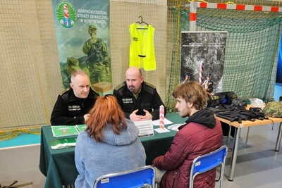 Funkcjonariusze Karpackiego Oddziału Straży granicznej rozmawiający z zainteresowanymi osobami służba w straży granicznej. na pierwszym planie dwie osoby  - kobieta i mężczyzna, którzy siedzą przed funkcjonariuszami. za funkcjonariuszami znajdują się banery informacyjne.