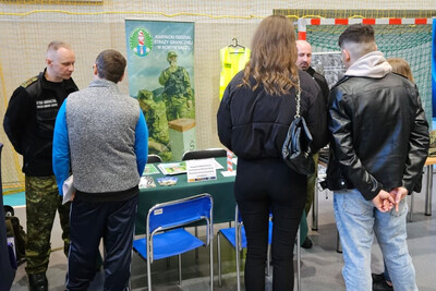 Funkcjonariusze Karpackiego Oddziału Straży granicznej rozmawiający z zainteresowanymi osobami służbą w straży granicznej. na pierwszym planie osoby, które stoją z funkcjonariuszami. za funkcjonariuszami znajdują się banery informacyjne.