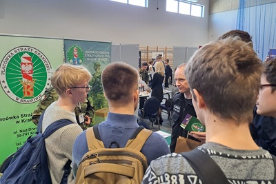 Funkcjonariusz Karpackiego Oddziału Straży granicznej rozmawiający z zainteresowanymi osobami służbą w straży granicznej. na pierwszym planie osoby, które stoją z funkcjonariuszem. za funkcjonariuszem znajdują się banery informacyjne.