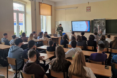 Funkcjonariuszka przemawia do zebranych uczniów podczas prelekcji. spotkanie odbywa się w szkolnej klasie. na pierwszym planie młodzież uczestnicząca w spotkaniu , na drugim planie funkcjonariuszka straży Granicznej omawiająca slajd, który jest wyświetlany.