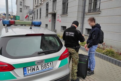 Uzbek nie wykonał decyzji, Mołdawianin przeterminował swój pobyt o 669 dni i był poszukiwany funkcjonariusz straży granicznej z zatrzymanym obywatelem Mołdawii który ma założone na ręce kajdanki. obok nich stoi bagaż zatrzymanego oraz pojazd służbowy straży granicznej.