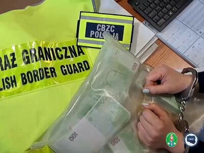 Na biurku leży odblaskowa  kamizelka z napisem Straż Graniczna, plakietka na rzepę z napisem  CBZC Policja, worek z wieloma banknotami w nominale 100 euro. na worku kobiece ręce zakute w kajdanki.  Obok po prawej klawiatura komputerowa i kalendarz. W prawym dolnym rogu, logotyp Karpackiego Oddziału Straży Granicznej i Centralnego Biura Zwalczania Cyberprzestępczości.