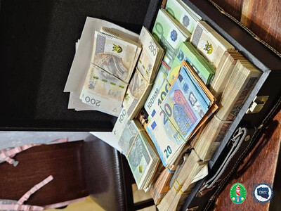 Na stole leży walizka z dużą ilością pieniędzy. Pliki banknotów spiętych gumkami o nominale 100 zł, 200 zł 100 euro i 20 euro. W prawym dolnym rogu, logotyp Karpackiego Oddziału Straży Granicznej i Centralnego Biura Zwalczania Cyberprzestępczości.
