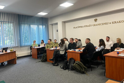 Szkolili Pograniczników z Karpackiego Oddziału Straży Granicznej funkcjonariusze, którzy są uczestnikami szkolenia. siedzą przy biurkach. nad nimi na ścianie znajduje się napis: Placówka straży granicznej w Krakowie oraz godło Polski.