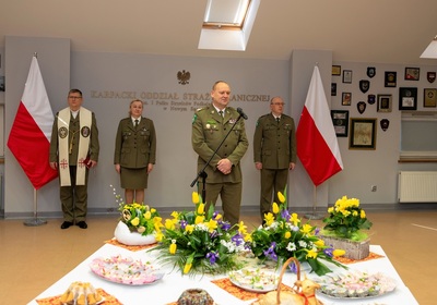 komendant Karpackiego Oddziału straży granicznej przemawiający do zebranych funkcjonariuszy i pracowników podczas spotkania świątecznego. Za komendantem stoją jego zastępcy oraz kapelan oddziału. Przed komendantem znajduje się nakryty stół do spotkania świątecznego z ozdobami świątecznymi oraz jajeczkami i babeczkami świątecznymi.