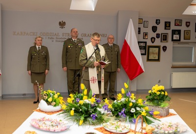 kapelan karpackiego oddziału straży granicznej odprawiający modlitwe i błogosławieństwo. za nim stoją komendant Karpackiego Oddziału straży granicznej oraz jego zastępcy Przed kapelanem znajduje się nakryty stół do spotkania świątecznego z ozdobami świątecznymi oraz jajeczkami i babeczkami świątecznymi. Wydarzenie odbywa się w Sali odpraw.