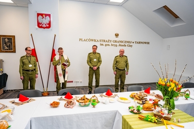 na pierwszym planie stoi zastępca komendanta Karpackiego Oddziału straży granicznej, obok niego stoi komendant placówki straży granicznej w Tarnowie oraz kapelan oddziału który błogosławi przygotowane posiłki i wszystkich zebranych podczas spotkania świątecznego, obok kapelana stoi zastępca komendant placówki straży granicznej w Tarnowie. Przed nimi znajduje się przygotowany stół do spotkania wielkanocnego na którym znajdują się talerze, ozdoby oraz dania świąteczne.