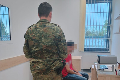 Rekordzista wśród nielegalnie przebywających cudzoziemców na terytorium RP zatrzymany mężczyzna siedzi na krześle w pomieszczeniu  służbowym straży granicznej. obok niego stoi funkcjonariusz straży granicznej który pilnuje zatrzymanego.