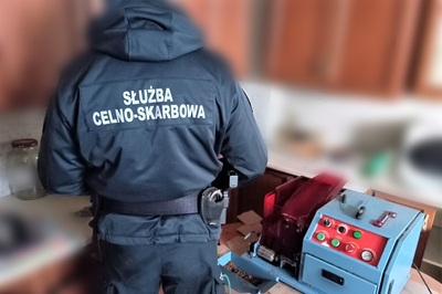 funkcjonariusz służby celno-skarbowej sprawdzający zatrzymany towar. obok maszyna prawdopodobnie do produkcji nielegalnego towaru. jest ona w kolorze niebieskim z czerwonymi wstawkami.